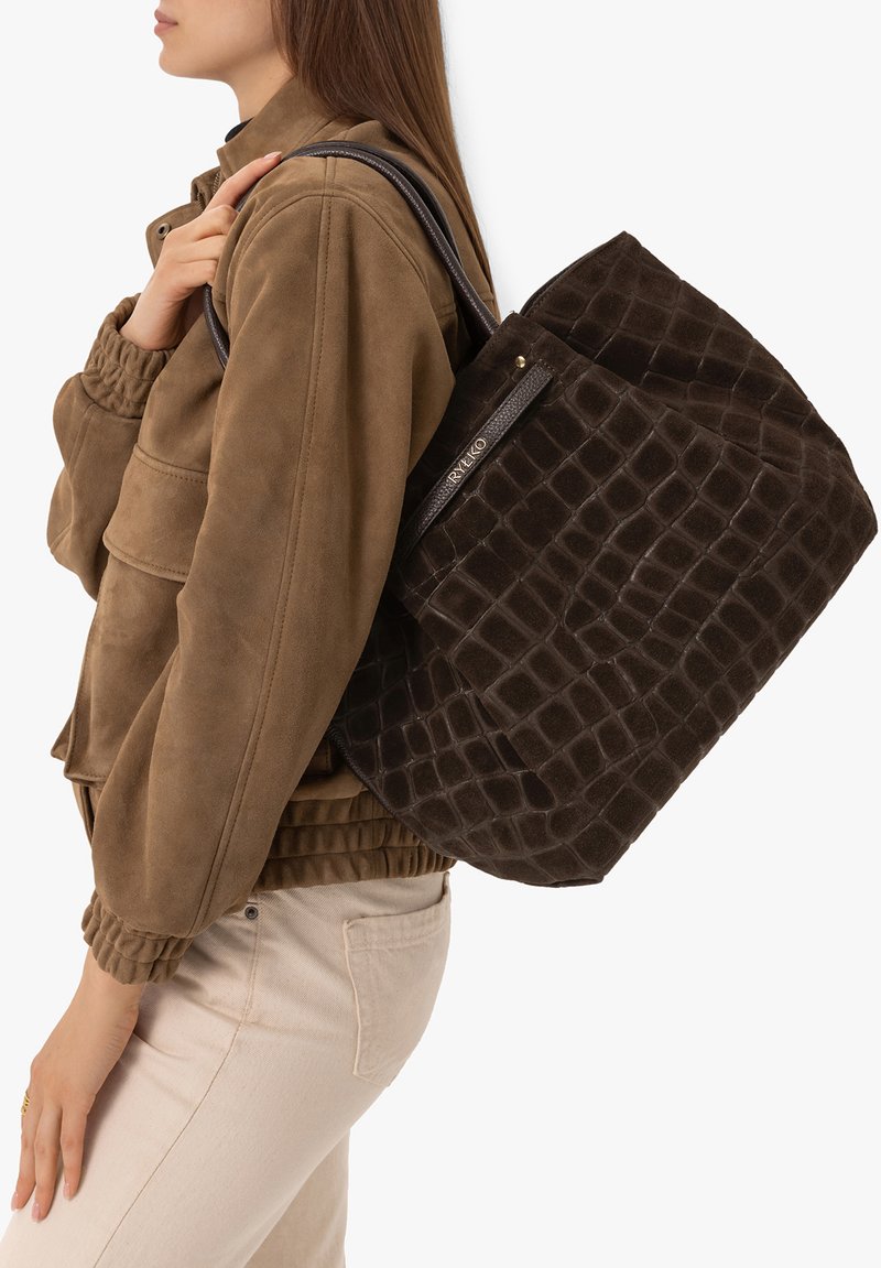 Femme portant une veste marron et un pantalon clair, transportant un grand sac cabas texturé marron foncé avec des bandoulières.