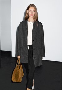 Szary, teksturowany płaszcz z czarnymi guzikami, noszony na białej bluzce i czarnych spodniach. Brązowa zamszowa torebka i białe buty dopełniają całość.