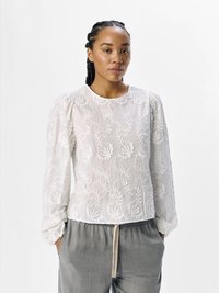 Blusa bianca ricamata con maniche a sbuffo e scollatura rotonda, caratterizzata da motivi floreali e una trama trasparente. Abbinata a pantaloni grigi.