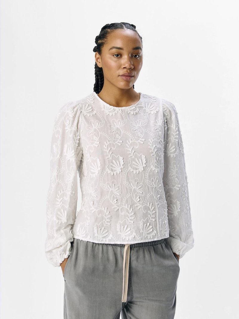Blusa bianca ricamata con maniche a sbuffo e scollatura rotonda, caratterizzata da motivi floreali e una trama trasparente. Abbinata a pantaloni grigi.