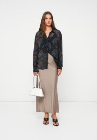 Blusa nera trasparente con fantasie floreali, gonna lunga beige, borsa bianca e sandali neri. Modella in piedi davanti a uno sfondo neutro.