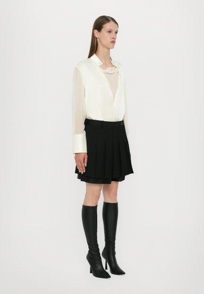 Camicia di seta bianca con scollatura profonda e maniche lunghe in raso, abbinata a una gonna nera plissettata e stivali neri con tacco a spillo fino al ginocchio.