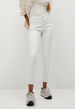 Person iført hvide, stribede, korte jeans, beige jakke, hvid top og grå sneakers, der står mod en ensartet lys baggrund.