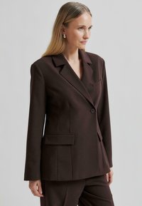 Hnědý přizpůsobený blazer s jedním knoflíkem, ostrými klopami a náprsní kapsou. Má hladkou strukturu a pevný siluetu.