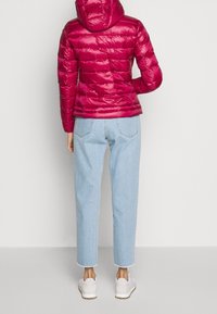 Doudoune fuchsia à capuche, design matelassé, associée à un jean bleu clair court et des baskets blanches, vue de dos.