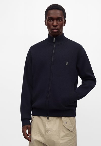 Homme portant un pull zippé bleu marine avec un petit logo sur la poitrine et un pantalon beige avec une poche boutonnée, debout devant un fond uni.