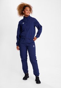 Mörkblå sportdräkt i lätt tyg. Innehåller en zip-jacka med elastiska ärmslut och byxor med elastisk midja. Försedd med en accentuerad logotyp.