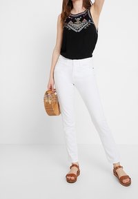Vrouw in een zwarte geborduurde mouwloze top, witte jeans, bruine sandalen en met een houten slatted handtas tegen een witte achtergrond.