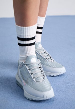 Jambes portant des chaussettes blanches avec deux bandes noires et des baskets athlétiques gris clair, debout sur un sol recouvert d’un tapis bleu.