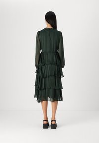 Grön lagerklänning med långa ärmar, volangkrage, genomskinligt tyg och ett flerlagers fåll. Har en figurnära midja och lätt textur.