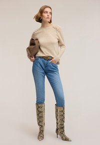 Béžový svetr s ozdobami na ramenou, světle modré džíny a vysoké boty s hadím vzorem na špičce. Hnědá psaníčko.