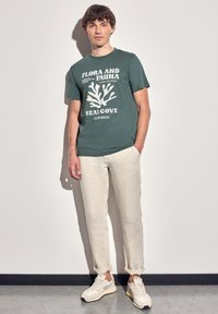 Tmavě zelené bavlněné tričko s bílou korálovou grafikou a textem, doplněné světle béžovými lněnými kalhotami. Tenisky jsou světlé s šedými akcenty.