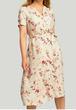 Vrouw draagt een beige midi-jurk met knopen aan de voorkant, korte mouwen en bloemmotief, met een strikceintuur in de taille, staand tegen een effen achtergrond.