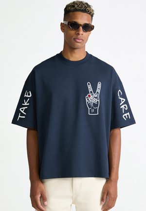 Homme portant un T-shirt bleu foncé oversize avec un graphisme de main de paix et le texte "TAKE" sur une manche et "CARE" sur l'autre, associé à des lunettes de soleil.