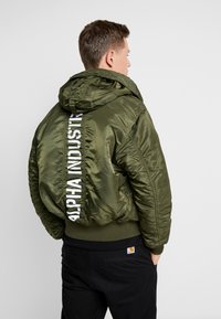 Maslinasto zelena bomber jakna s sjajnim završetkom, kapuljačom i bijelim tiskanim logotipom na leđima. Ima rebraste rukave i donji rub.