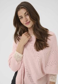 Šviesiai rožinė pončo su zigzagų raštais, pagaminta iš minkštos medžiagos. Yra nėrinių apdaila rankovėse, suteikianti jaukų ir tekstūruotą išvaizdą.