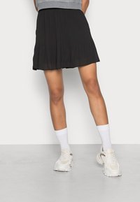 Persoon in een zwarte geplooide rok, witte sokken en witte chunky sneakers, staand tegen een effen lichte achtergrond.