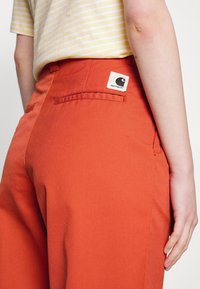 Oranje broek gemaakt van stevig materiaal, met een achterzak met een merklabel, en een getailleerde pasvorm die wordt benadrukt door nauwkeurige stiksels.