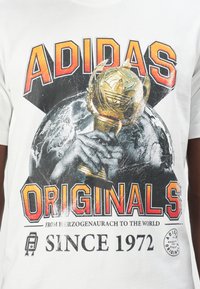 Hvid bomulds T-shirt med et grafisk print af et trofæ og en globus. Fed "ADIDAS ORIGINALS" tekst i rød og sort med slidt detaljer.