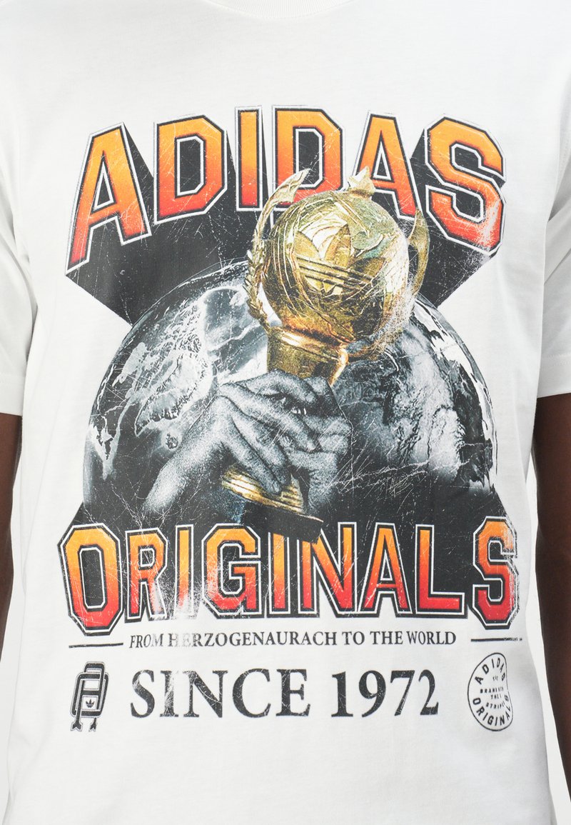 Camiseta de algodón blanca con un gráfico de un trofeo y un globo. Texto en negrita "ADIDAS ORIGINALS" en rojo y negro, con un aspecto desgastado.