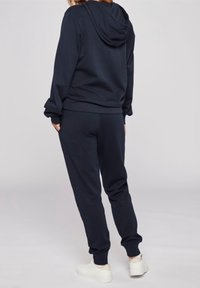 Námořnická mikina s kapucí a volným střihem, žebrovanými manžetami a odpovídajícími tepláky. Vyrobena z měkké látky s minimalistickým designem. Bílé tenisky dokončují vzhled.