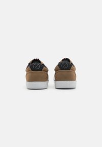 Baskets en daim marron avec des accents noirs, semelles en caoutchouc blanches et talons arrondis. Dotées d'un logo circulaire sur le talon.