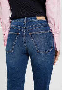 Vaqueros de mezclilla azul oscuro con un diseño ajustado, que presentan dos bolsillos traseros, costuras sutiles y un parche de cuero marrón de la marca. Textura mate.