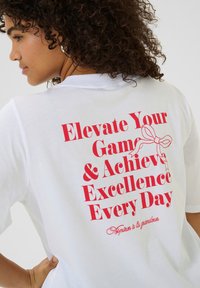 T-shirt en coton blanc présentant un texte rouge audacieux au dos : "Élevez Votre Jeu & Atteignez l'Excellence Chaque Jour" avec un graphique de ruban.