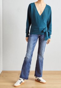 Teal geribbelde wikkeltop met een diepe V-hals en strikdetail bij de taille, gecombineerd met lichtblauwe flared jeans en witte sportschoenen.