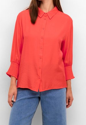 Kvinde iført koralfarvet skjorte med knapper og let opsamlede 3/4-ærmer samt blå jeans, stående foran en ensfarvet lys baggrund.