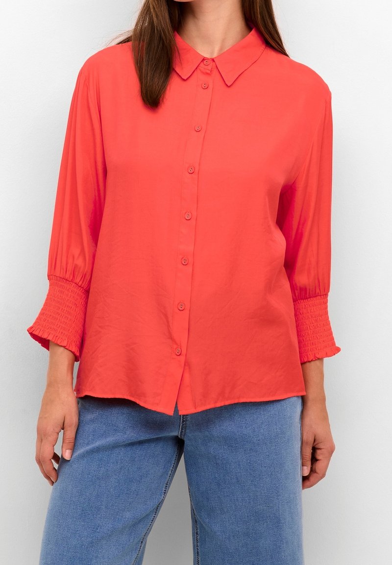Femme portant un chemisier corail boutonné avec des manches 3/4 froncées et un jean bleu, debout devant un fond clair uni.