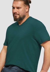 Dunkelgrünes V-Ausschnitt-T-Shirt aus glattem Stoff, kurze Ärmel und ein dezentes Logo auf der Brust. Lockere Passform mit klassischem Design.