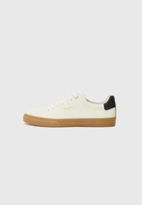 Zapatilla de lona blanca con suela de goma, acento negro en el talón, punta redonda y diseño con cordones. Presenta el logo de la marca en dorado en el lateral.