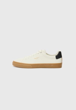 White canvas sneaker with a gum sole, black heel accent, round toe, and lace-up design. Features brand logo in gold on the side.