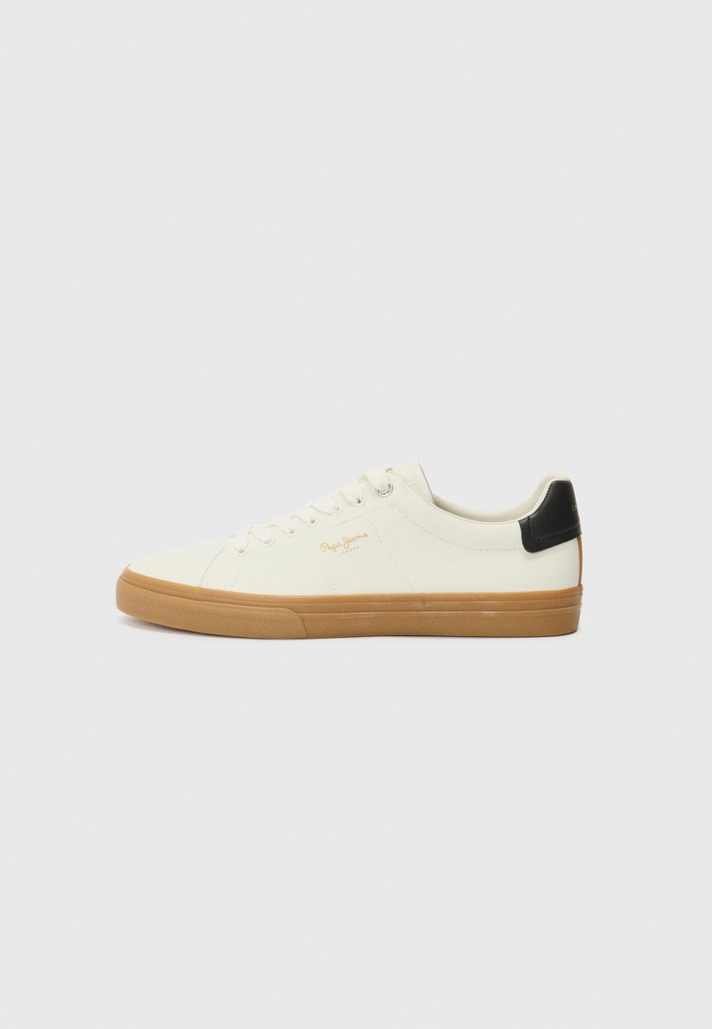Zapatilla de lona blanca con suela de goma, acento negro en el talón, punta redonda y diseño con cordones. Presenta el logo de la marca en dorado en el lateral.