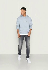 Hanorac albastru deschis cu șnururi, asortat cu blugi gri închis și adidași albi. Hanoracul are un buzunar tip kangaroo și manșete din tricot.