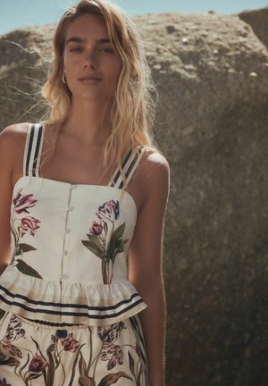 Junge Frau mit langen blonden Haaren, die ein ärmelloses weißes Oberteil mit Blumenmuster und einen passenden Rock trägt und im Freien in der Nähe großer Felsen steht.