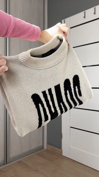 Ljusbeige tröja med ribbad halsringning och en djärv svart textdesign. Stickningsstrukturen är synlig och hålls i en hand mot en neutral bakgrund.