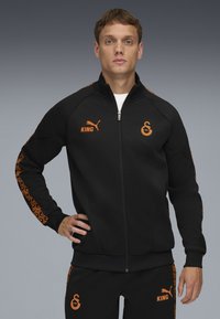 Schwarze Jacke aus synthetischem Stoff, mit orangefarbenen Akzenten, seitlichem Reißverschluss und tribalem Muster an den Ärmeln; Logo auf der Brust und den Hosen.