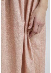 Tessuto di abito rosa chiaro con un delicato motivo floreale e paisley. Mano visibile che afferra una piega, mostrando la morbidezza della texture e la caduta.