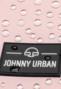 Fekete gumi címke fehér domború felirattal "JOHNNY URBAN" szöveggel, amely egy texturált rózsaszín szövet háttéren helyezkedik el, látható vízcseppekkel.