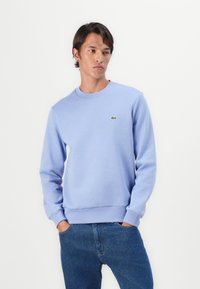 Lyseblå fl fleece sweatshirt med rund halsudskæring, ribbede ærmer og kant. Har et lille grønt logo på venstre bryst. Bæret med blå jeans.
