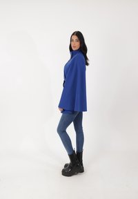 Capa azul con cuello alto, tejido texturizado, combinada con jeans de cintura ajustada y botas negras gruesas. Diseño sencillo, sin patrones visibles.