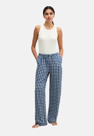 Vrouw staat blootsvoets, draagt een wit mouwloos topje en losse blauw-wit geruite broek met handen in de zakken.