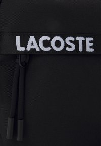 Zaino in tessuto nero con una finitura testurizzata, caratterizzato da un logo "LACOSTE" ricamato in bianco e accenti in nero con coulisse.