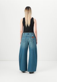 Femme aux longs cheveux blonds portant un haut sans manches noir et un jean bleu à jambes larges, se tenant dos à un fond blanc.