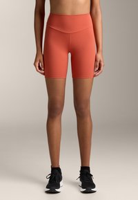 Magas derekú, rozsdabarna rövidnadrág rugalmas anyagból, sima textúrával és combközép hosszúsággal, fekete sportcipővel párosítva.