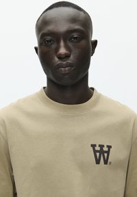 Felpa beige realizzata in tessuto morbido, con colletto rotondo e un logo grafico "W" marrone scuro sul lato sinistro del petto.