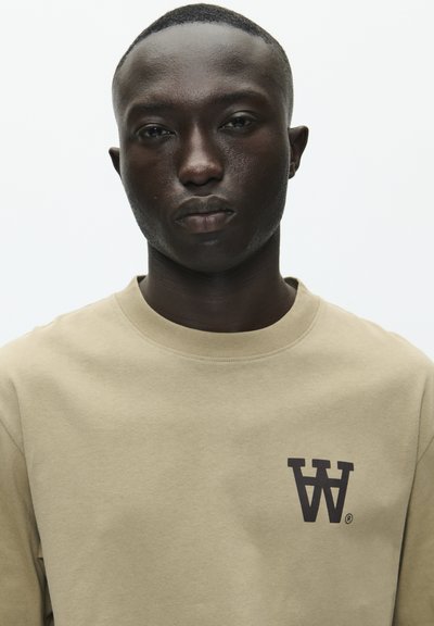 Sweatshirt beige en tissu doux, avec un col rond et un logo graphique "W" marron foncé sur la partie gauche de la poitrine.