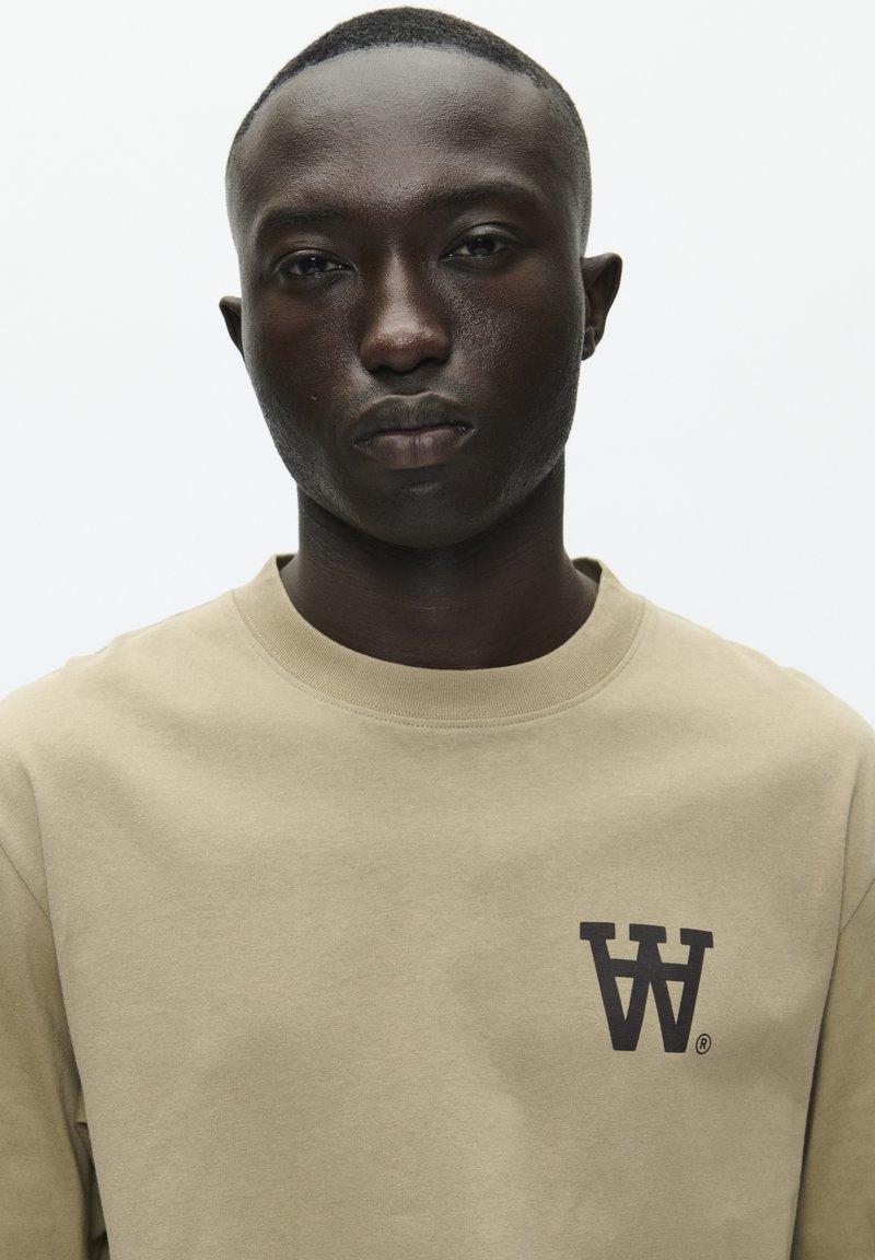 Sudadera beige hecha de un tejido suave, con cuello redondo y un logo gráfico en color marrón oscuro con la letra "W" en la parte izquierda del pecho.